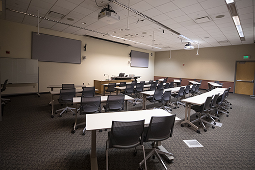 Photo of Doyle 145 - larger classroom with 2 large screens, a lectern, and tables and chairs facing the front