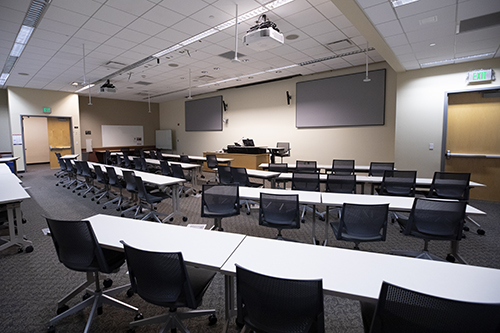 Photo of Doyle 146 - larger classroom with screens, a lectern, tables, chairs, whiteboards, video conferencing